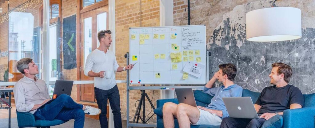 Team brainstorming in a bright brick office: presenter at a whiteboard with sticky notes, others watching.