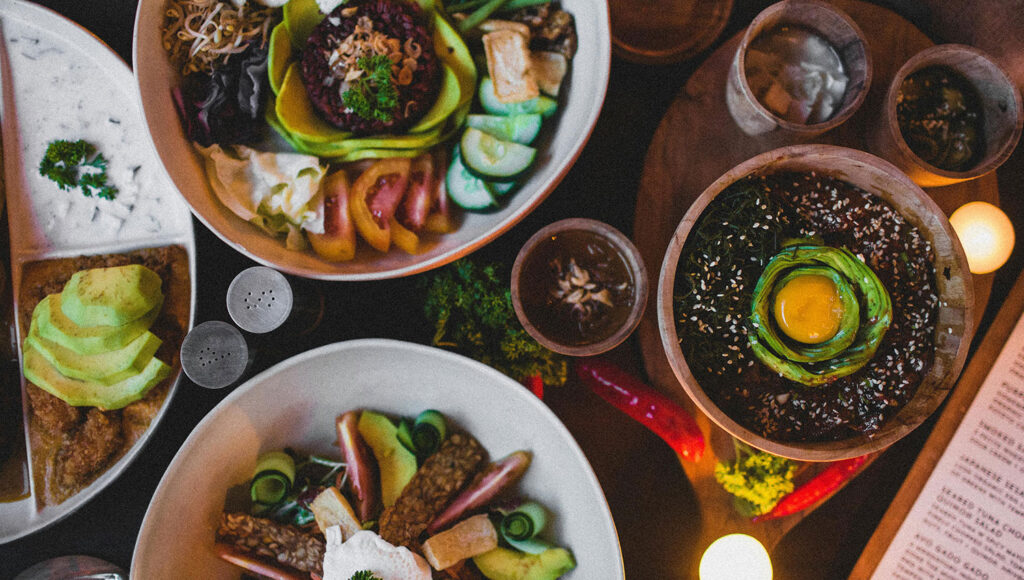 Colorful meal spread on a dark table: avocado toast, veggie bowls, and a sesame rice bowl topped with avocado and egg rosette.