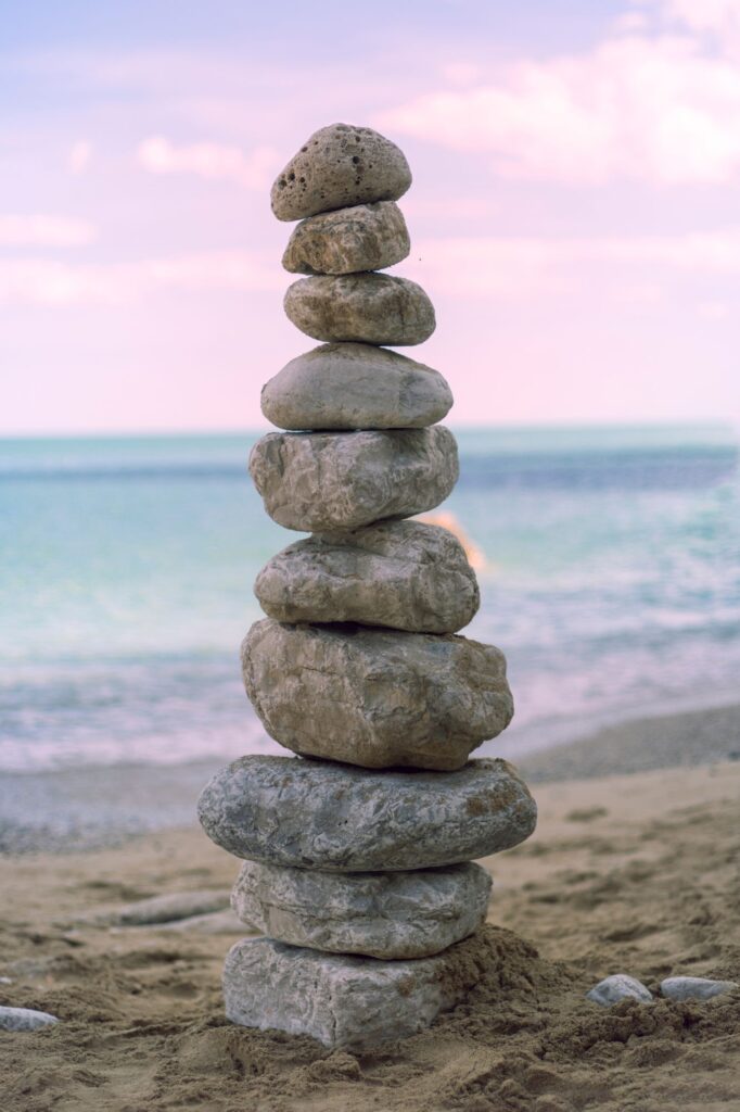 Seven stacked rocks balanced on a rocky base on a sandy beach with a calm sea and pastel sky in the background by the shore at sunset/sunrise.