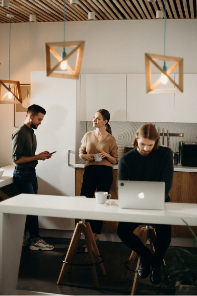 Three coworkers in a modern kitchen-like workspace: man on left using a phone, woman center with a mug, man right at a laptop; geometric pendant lights above.