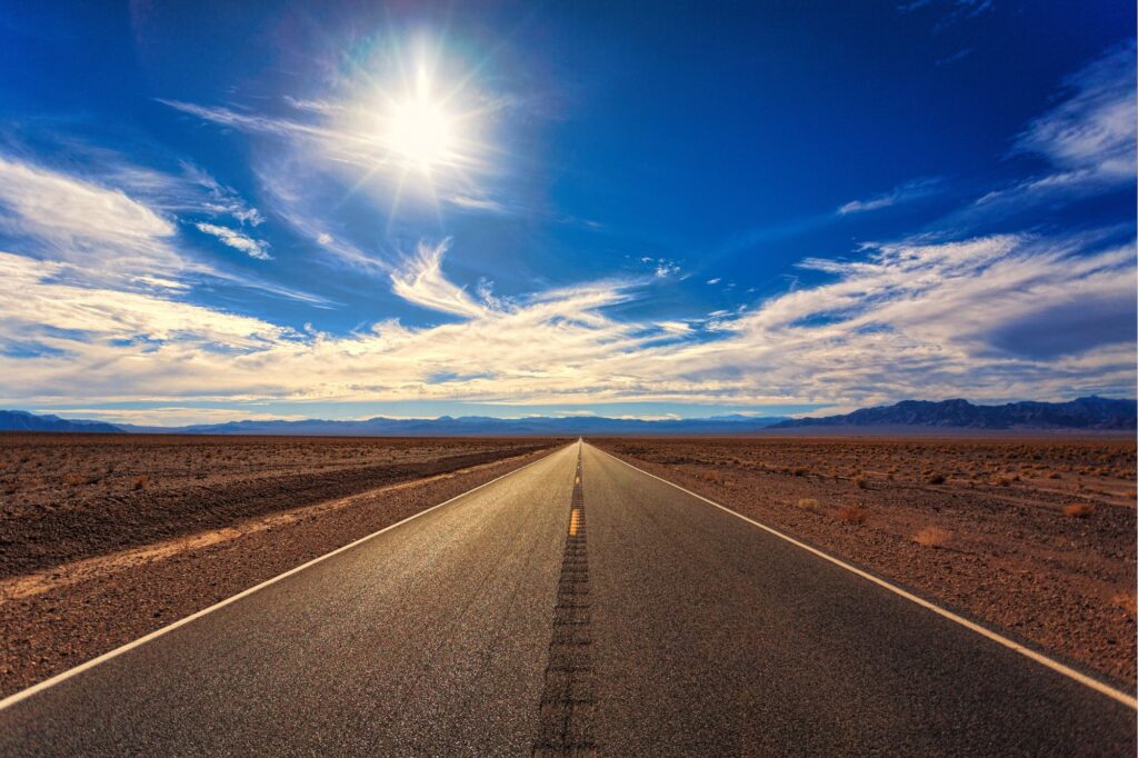 Long straight desert highway stretching to the horizon under a bright sun and blue sky with distant mountains.