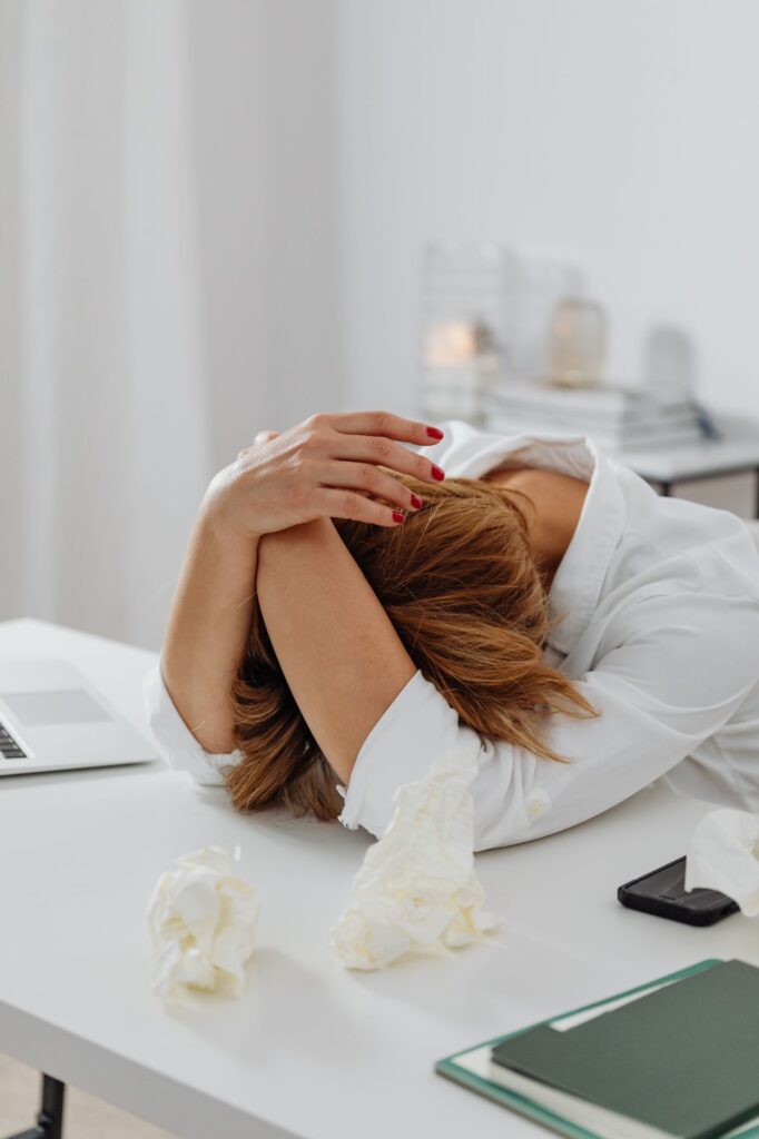 Person resting head on desk with hands clasped, surrounded by crumpled papers and a laptop, suggesting stress or fatigue in a workspace