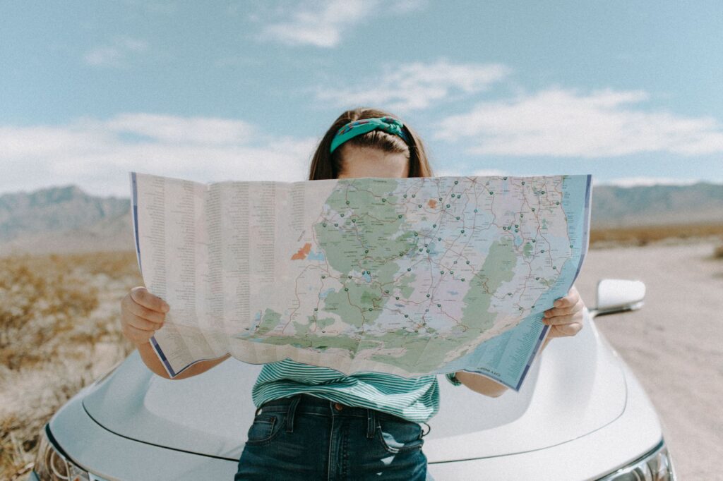 Person holding a large folded map outdoors beside a light blue car in a desert landscape, wearing a teal headband and striped shirt.