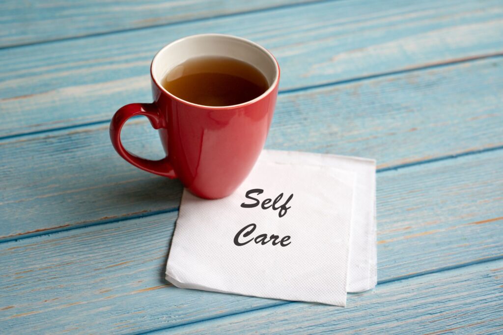 Red ceramic mug filled with tea sits on a light blue, weathered wooden surface beside a napkin that reads 'Self Care'.