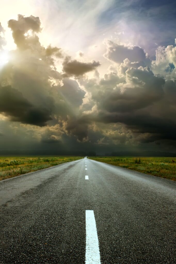 Long straight road disappearing into a stormy horizon under dramatic clouds, with open fields on either side.
