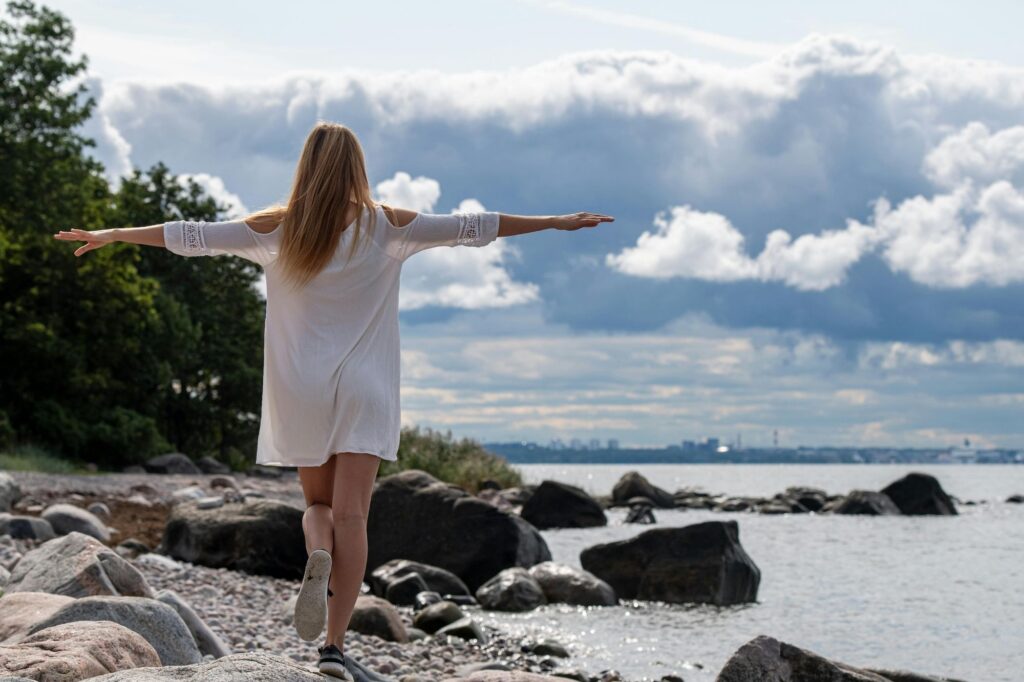 Woman in a white dress stands on rocky shoreline with arms outstretched, facing the water and cloudy sky.