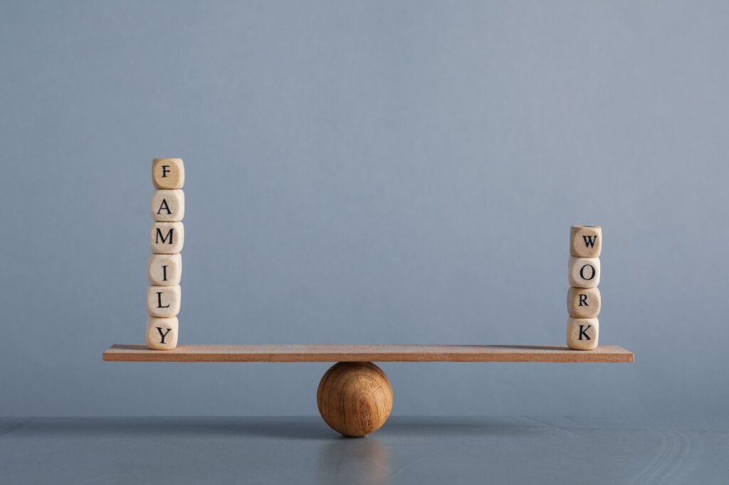 Wooden balance scale with a tall stack spelling 'FAMILY' on the left and a shorter 'WORK' stack on the right, showing family–work balance.