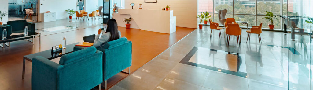 Modern office lobby with teal sofa, a person seated on the phone, orange chairs, and a white reception desk near glass walls with potted plants.