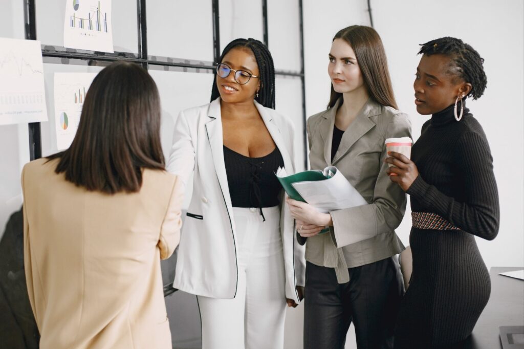 Four professionals in a meeting discuss charts and graphs posted on a wall-mounted board in a bright office.