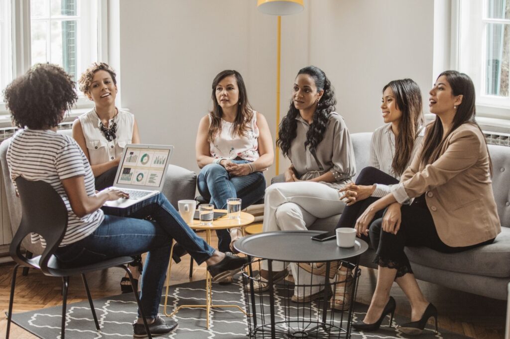 Six women of diverse backgrounds sit in a bright living-room, forming a loose circle and discussing, with a laptop showing charts nearby.
