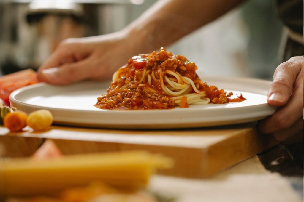 Plate of spaghetti with meat sauce on a white dish, two hands nearby as if serving or about to eat.