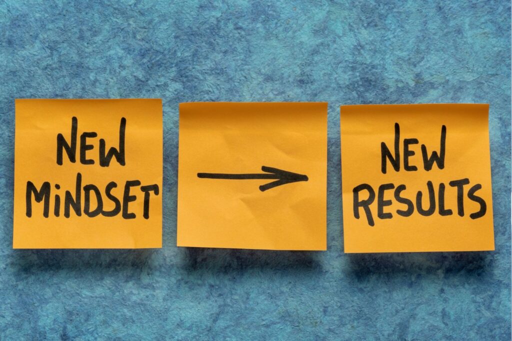 Three yellow sticky notes on a blue textured surface: left note reads 'New Mindset', middle note shows a rightward arrow, right note reads 'New Results'.