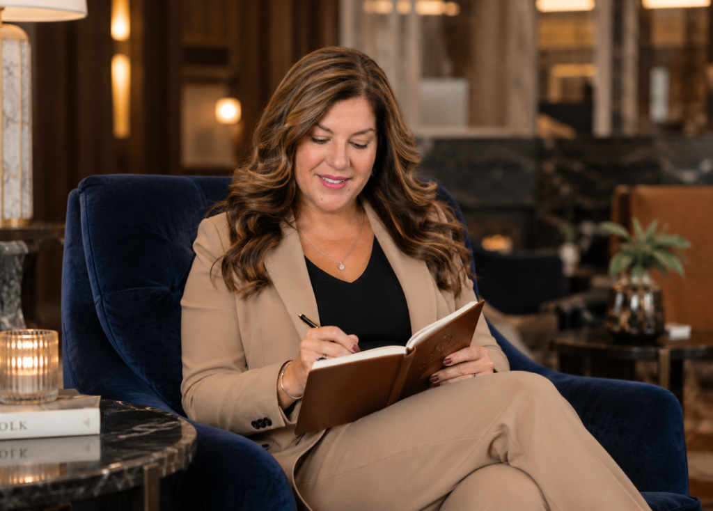 Woman in a tan suit sits in a deep blue velvet chair, writing in a notebook in a warm, stylish lounge.