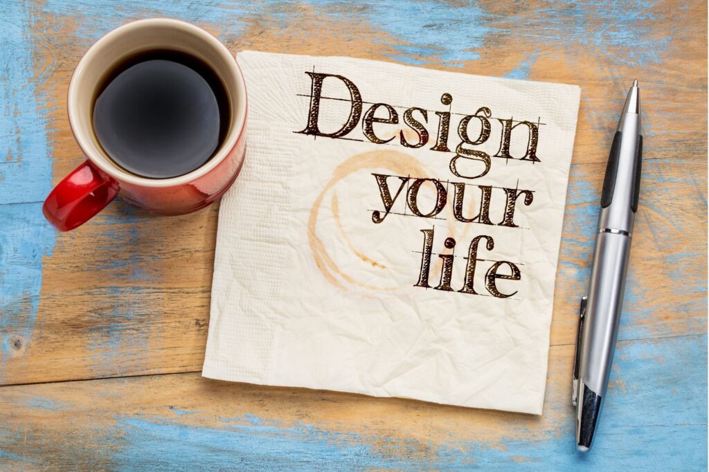 Red mug of coffee beside a napkin that reads 'Design your life' and a silver pen on a blue-washed wooden table.