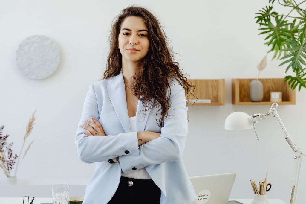 Professional woman standing with arms crossed in a bright office, wearing a light blue blazer.