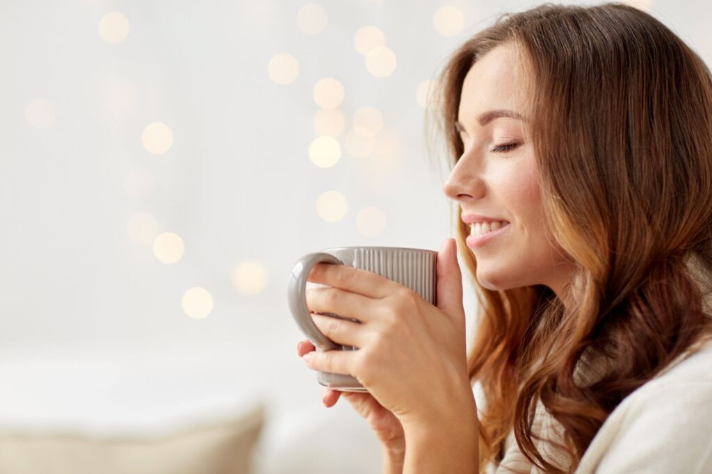 Woman with long brown hair smiling with eyes closed while holding a gray mug near her lips; warm bokeh lights in the background.