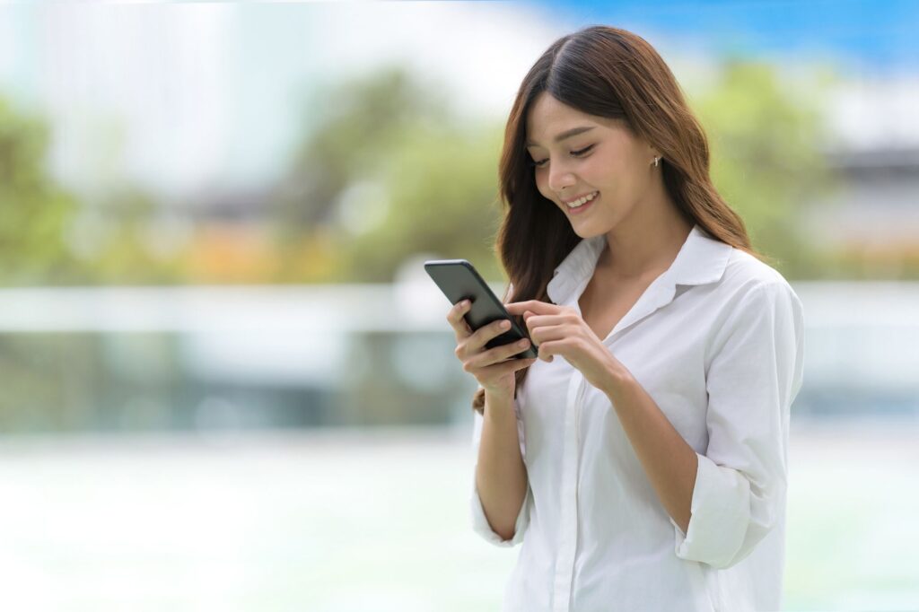Woman in a white shirt looking down at a smartphone outdoors, smiling slightly in a bright, blurred background