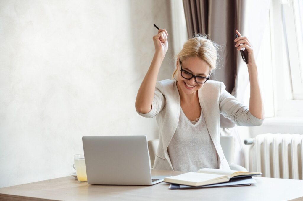 Woman with glasses sits at a laptop on a wooden desk, smiling and raising her arms in triumph.