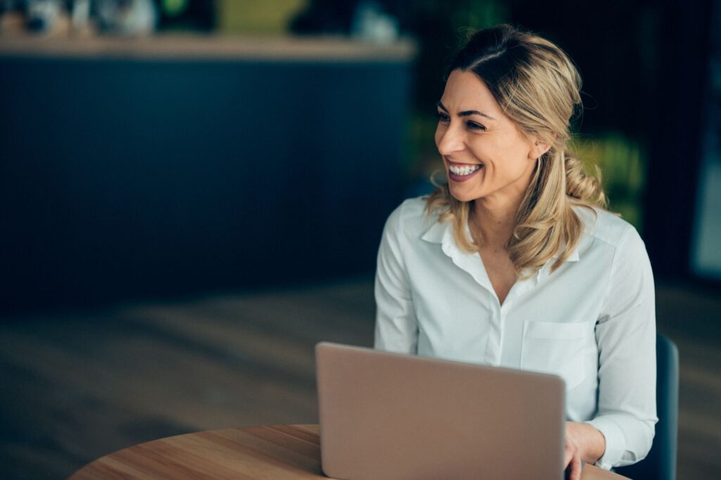 Smiling woman in a white shirt sits at a wooden table with a laptop in front of her.