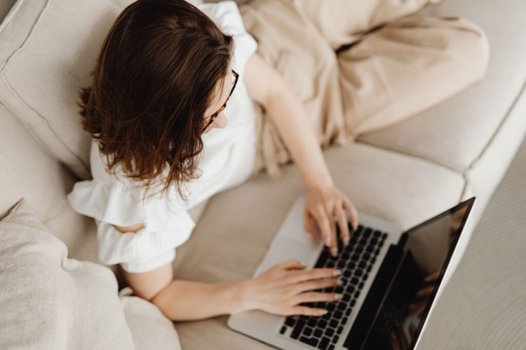 Overhead view of a woman with brown hair and tortoiseshell glasses typing on a laptop while seated on a beige sofa.