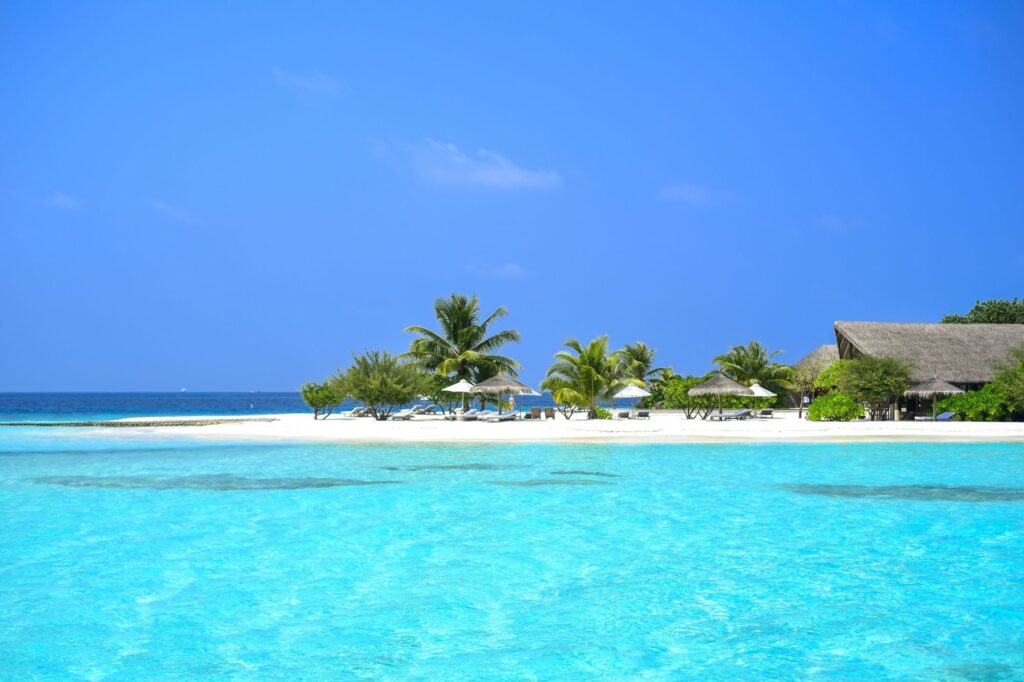 Tropical beach scene with turquoise water, white sand, palm trees and thatched-roof huts under a clear blue sky.