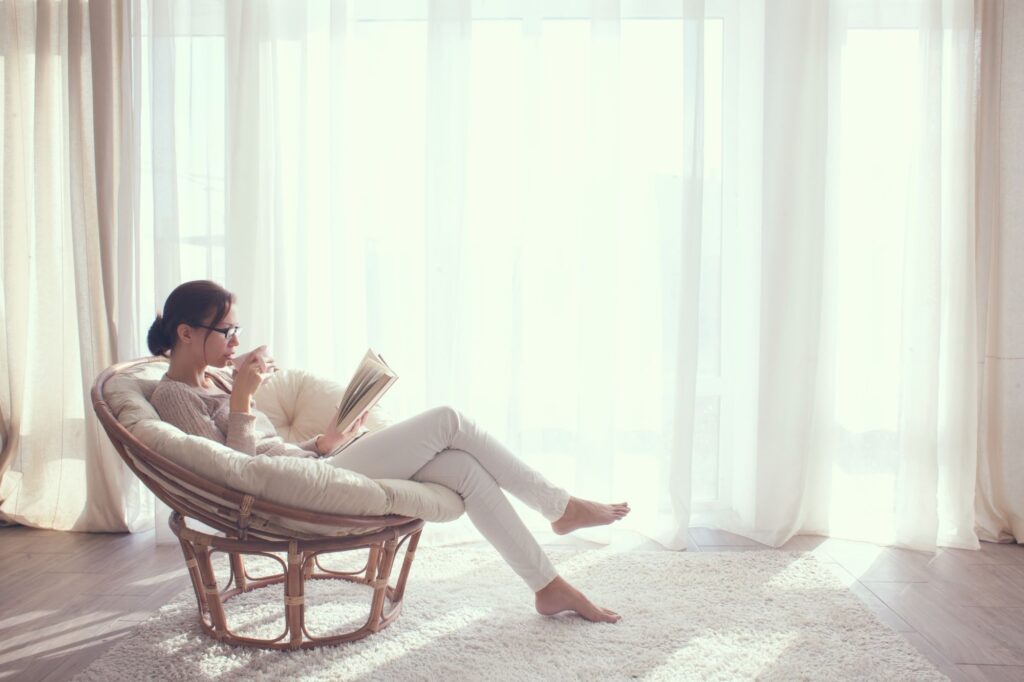 Woman lounging in a wicker chair reading a book by a bright window with sheer curtains.