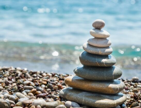 Stack of smooth, rounded stones balanced into a small cairn on a pebbled beach with the blue ocean in the background.