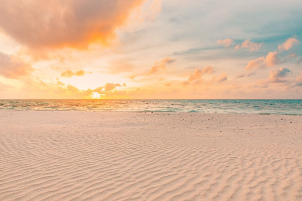 Sunset over a calm ocean with a pink-orange sky and rippled sandy shore in the foreground.
