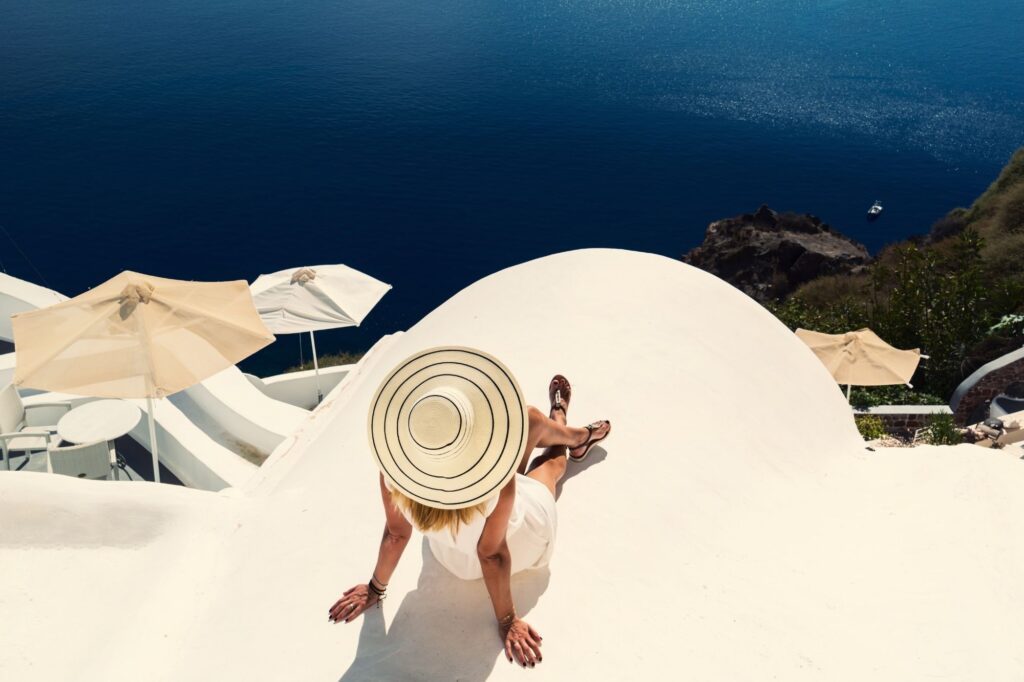 Person lounging on a white terrace with a wide-brimmed hat, looking toward deep blue sea with beige umbrellas nearby