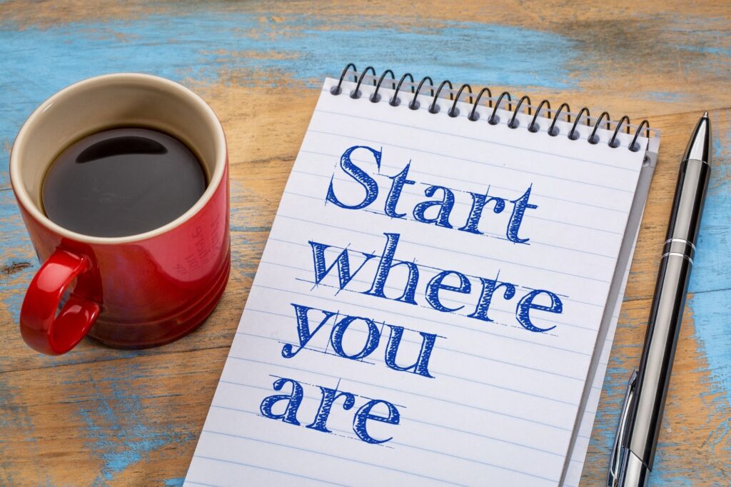 Red mug of coffee beside a spiral notebook reading 'Start where you are' on a rustic blue wooden table with a pen nearby