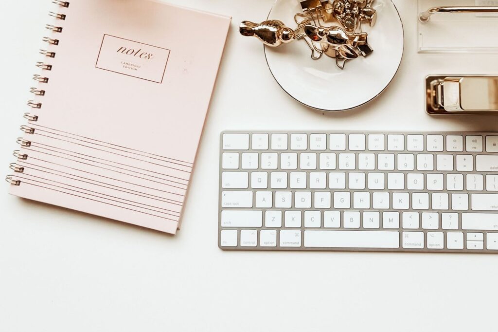 Top-down view of a white keyboard on a desk beside a pink spiral notebook and a plate filled with silver binder clips and other metal office items.
