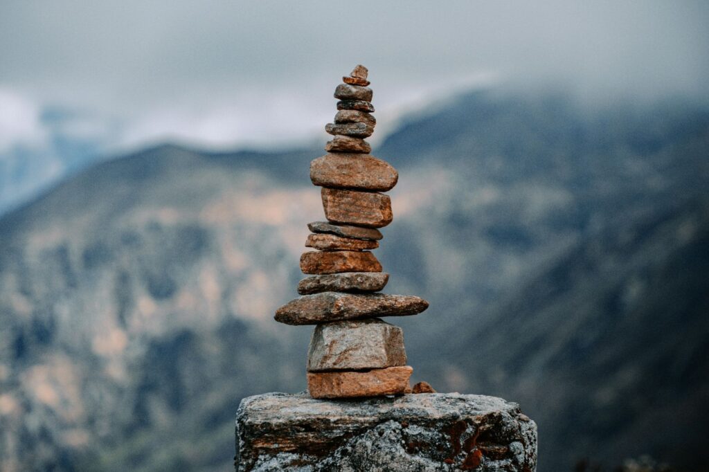 Tall cairn of stacked stones balanced on a rocky pedestal, with a misty mountain backdrop behind it.