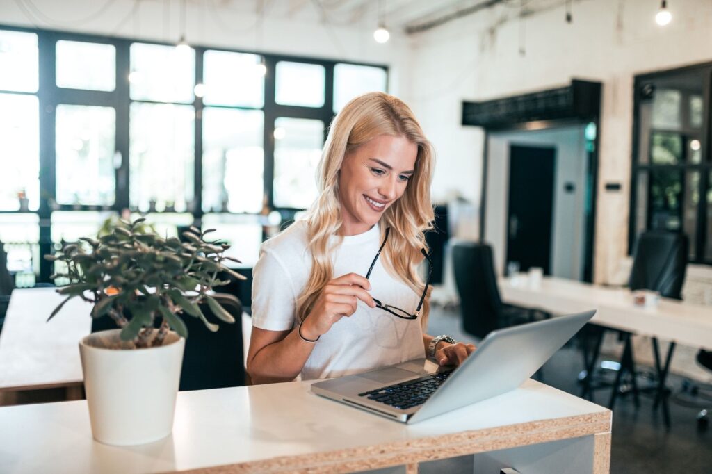 Smiling woman in a white shirt working on a laptop at a bright office desk with a plant nearby.