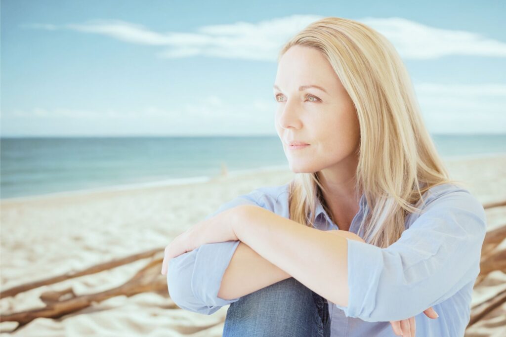 Woman sitting on a sandy beach, looking thoughtfully toward the sea on a sunny day.