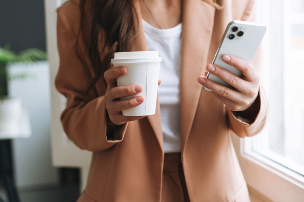 Person wearing a beige coat holding a white disposable coffee cup in one hand and a smartphone in the other indoors by a window.