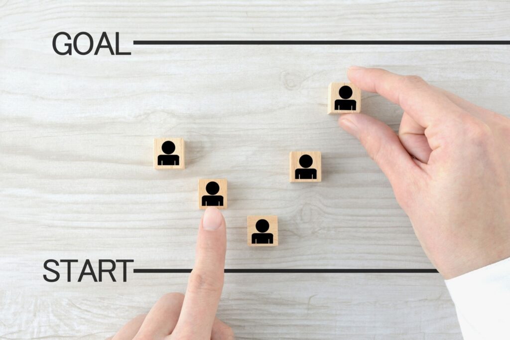 Hands place wooden blocks with person icons along a start-to-goal path on a light wood surface, illustrating goal setting and progression.