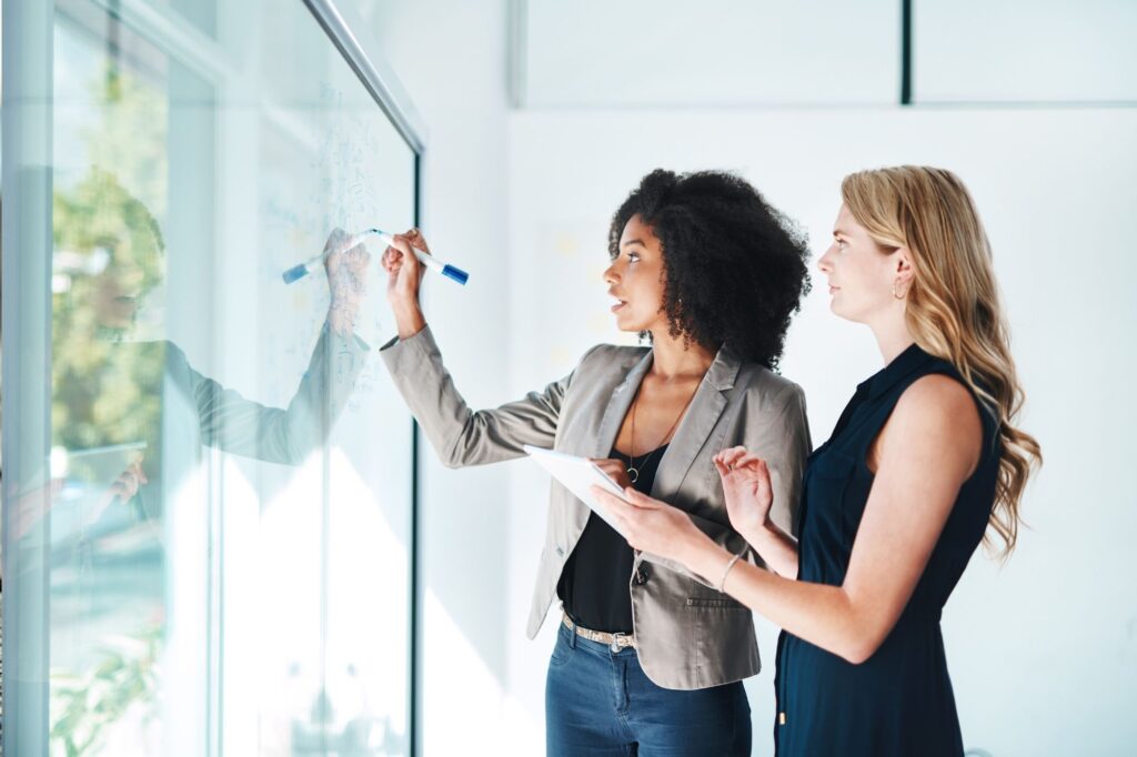 Two women colleagues collaborate at a glass whiteboard, one writes with a marker while the other holds a notepad or papers.