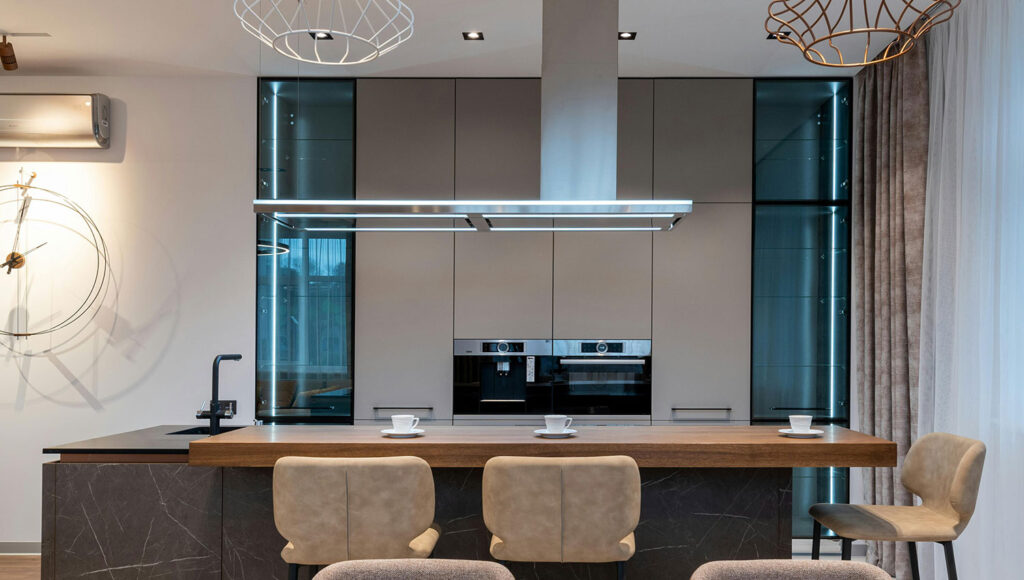 Modern open-plan kitchen with a dark marble island, tall grey cabinets, and built-in ovens in the center. Three beige chairs face the island.