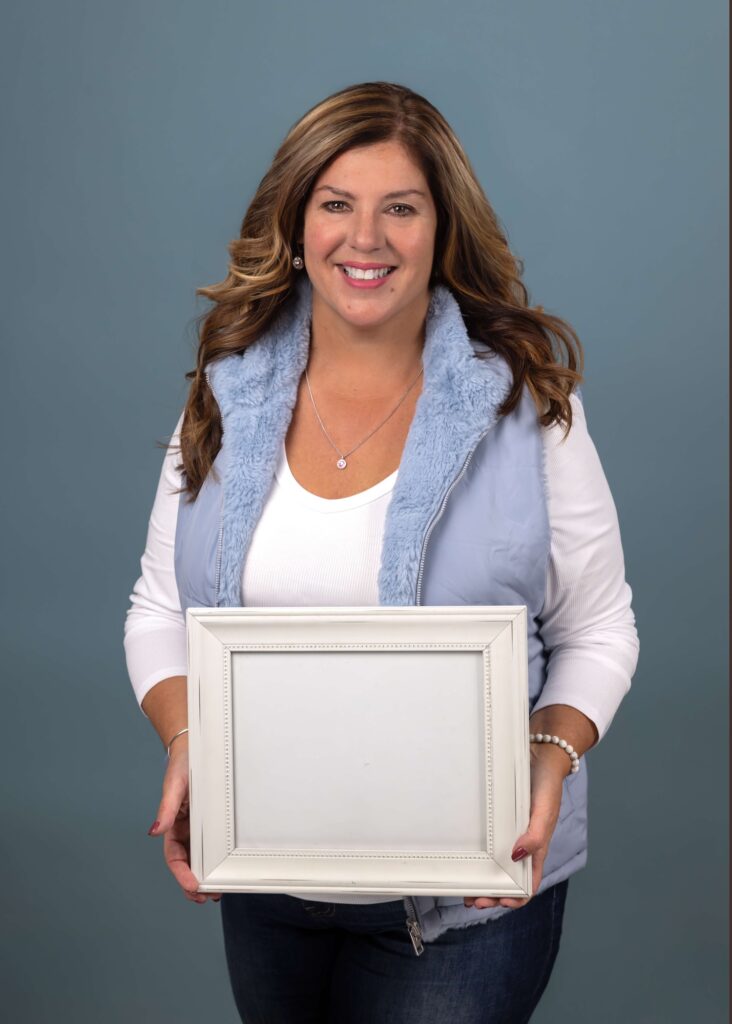 Smiling woman in a light blue vest holds a blank white picture frame in front of her against a blue background.